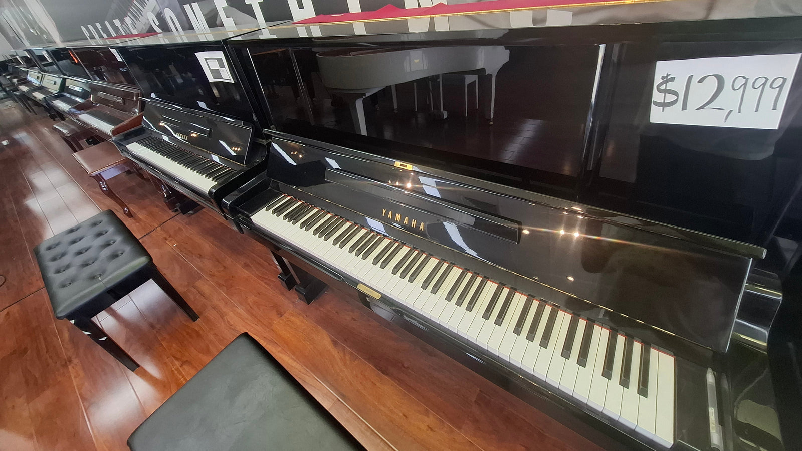 A Yamaha upright piano in glossy black, shown in a showroom with other pianos, wooden floors, and a price tag reading $12,999 attached to the open lid.